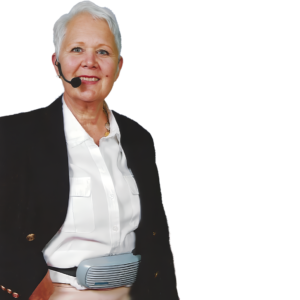 An older woman with short gray hair wears a ChatterVOX® Pro w/headset mic and a black blazer over a white shirt, smiling as she stands against a plain white background, with the device secured around her waist.