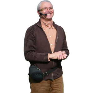 An older man with gray hair and glasses smiles while wearing a ChatterVOX® Model 6 Coaching Amplifier headset and speaker around his waist, dressed in a brown zip-up sweater over a striped shirt.
