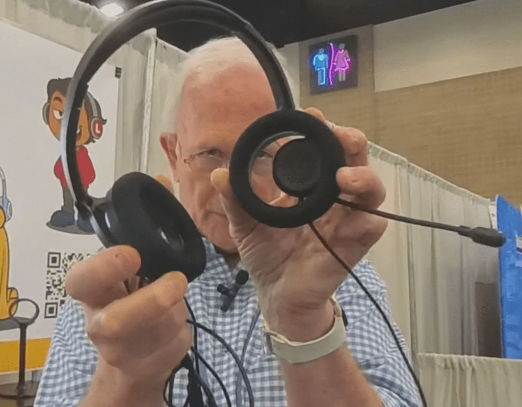 An older man with white hair holds a black headset with a microphone toward the camera at a convention or expo, showcasing Headphones for schools. Booths, a cartoon poster, and restroom signs can be seen in the background.
